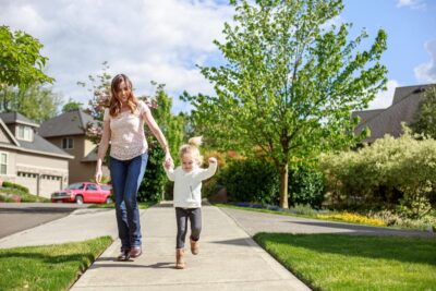 A mother and young child walking hand-in-hand along a sunny suburban sidewalk
